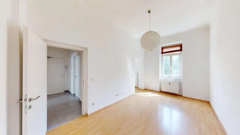 Helles, leeres Zimmer mit Parkettboden und einem Fenster, das viel Tageslicht hereinlässt.
