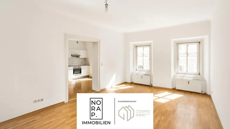Helles Zimmer mit Parkettboden und weißen Wänden, das zu einer Küche führt. Sonnenlicht fällt durch die Fenster.