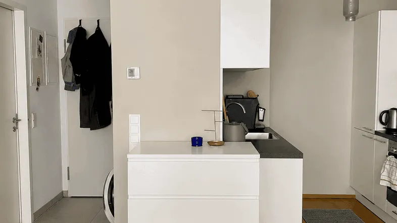 Moderne Küchenzeile mit weißen Schränken und dunkler Arbeitsplatte, offen zum Eingangsbereich mit Waschmaschine.