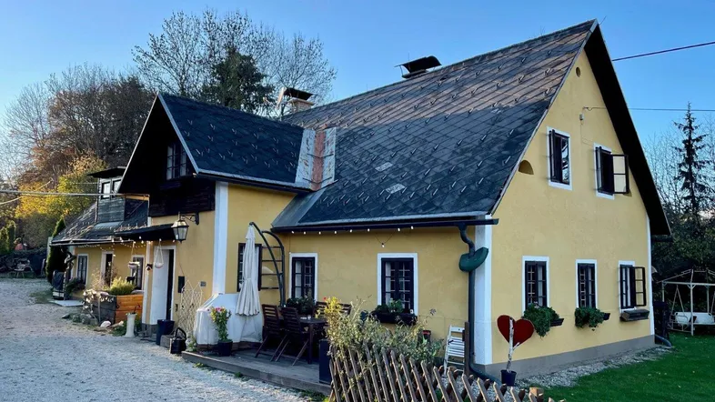 Gelbes Landhaus mit dunklem Dach, kleinen Fenstern und einer kleinen Terrasse im Freien.