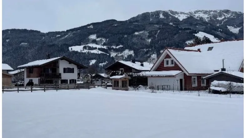 Winterliche Landschaft mit schneebedeckten Häusern und Bergen im Hintergrund unter bewölktem Himmel.