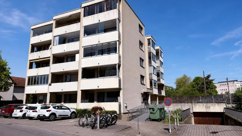 Modernes Mehrfamilienhaus mit Balkonen, Tiefgaragenzufahrt und Parkplätzen im Freien unter blauem Himmel.