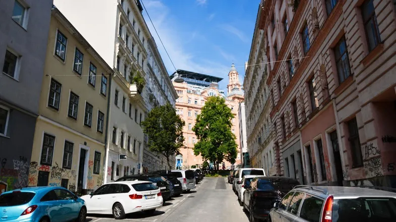 Blick auf eine belebte Stadtstraße mit geparkten Autos und hohen Wohngebäuden unter blauem Himmel.