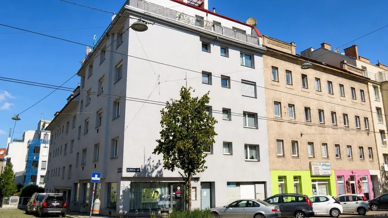 Mehrstöckiges Wohngebäude an einer Straße mit geparkten Autos und Geschäften im Erdgeschoss.