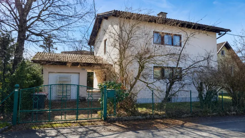 Einfamilienhaus mit Satteldach, umgeben von Bäumen und einem grünen Zaun im Vorgarten.