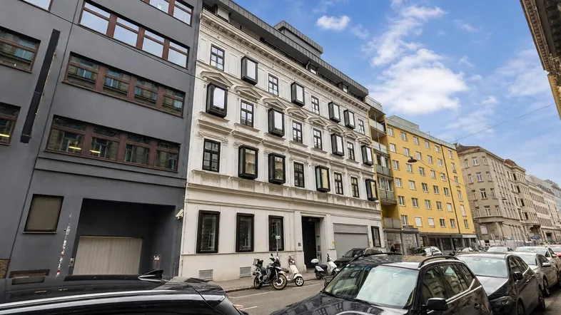 Blick auf eine belebte Straße mit verschiedenen Gebäuden, geparkten Autos und einem Roller in der Stadt.