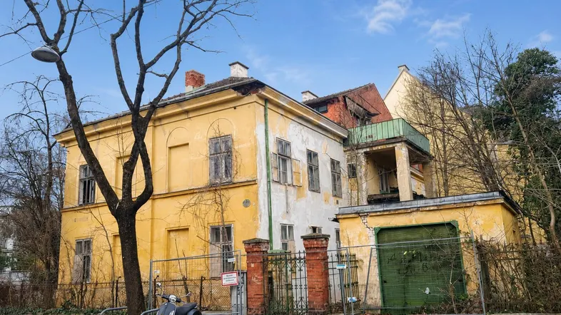 Altes, verfallenes Gebäude mit gelber Fassade und kahlen Bäumen, das auf eine Sanierung wartet.