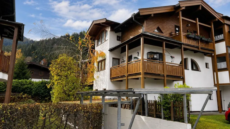 Mehrstöckiges Gebäude im alpinen Stil mit Holzbalkonen und einem Carport im Vordergrund, umgeben von grüner Natur.