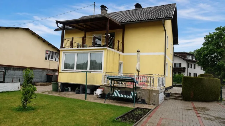 Gelbes Einfamilienhaus mit Balkon, kleiner Terrasse und gepflegtem Gartenbereich unter blauem Himmel.