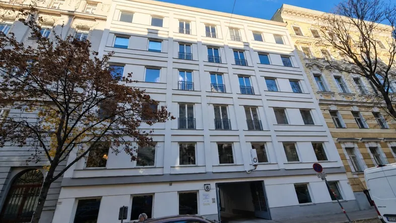 Mehrstöckiges Wohngebäude mit weißer Fassade und vielen Fenstern, davor ein geparktes Auto.