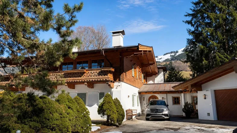 Traditionelles Alpenhaus mit Holzbalkonen, weißer Fassade und Bergblick, umgeben von Bäumen.