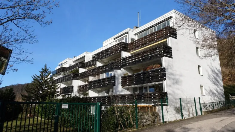 Mehrfamilienhaus mit zahlreichen Balkonen und umgebender Natur unter blauem Himmel.