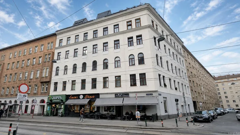 Mehrstöckiges Gebäude in urbaner Umgebung mit Geschäften im Erdgeschoss und geparkten Autos auf der Straße.