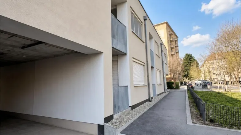 Moderne Gebäudefassade mit Tiefgaragenzufahrt, Balkonen und Rollläden unter blauem Himmel.
