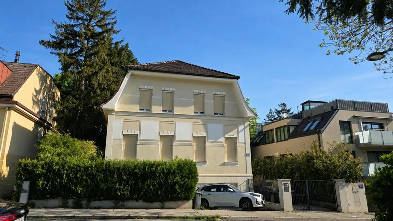 Elegantes Wohnhaus mit heller Fassade, Mansarddach und geschlossenen Fensterläden, umgeben von gepflegten Hecken und Bäumen.