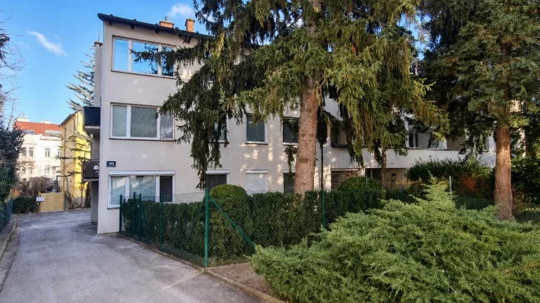 Mehrfamilienhaus mit grüner Umgebung und Zufahrt, umgeben von hohen Bäumen.