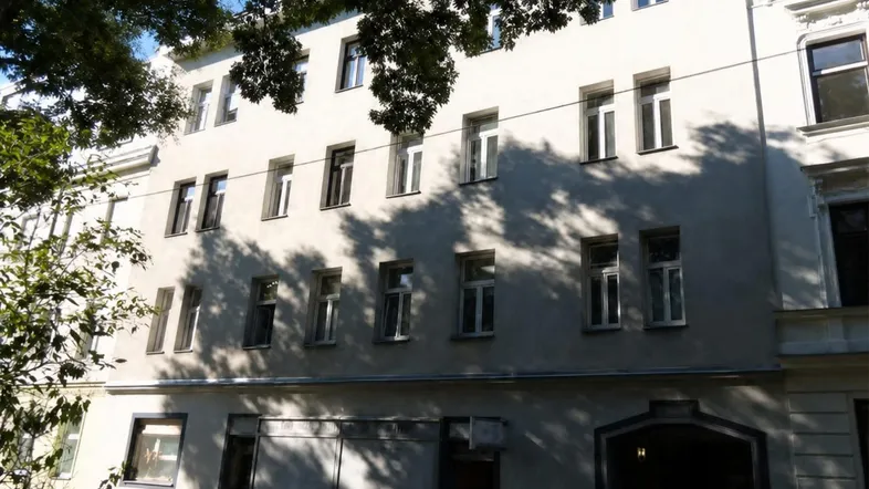 Fassade eines Mehrfamilienhauses mit Bäumen und geparkten Motorrollern im Vordergrund, bei Sonnenschein.