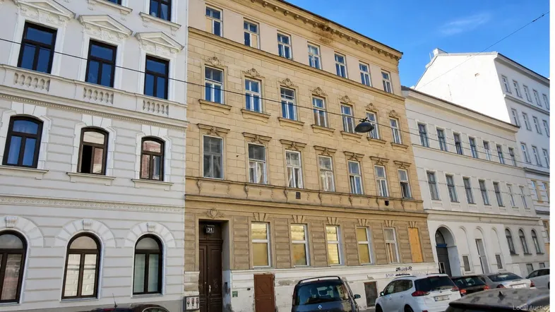 Historische Fassade eines Mehrfamilienhauses in einer städtischen Umgebung mit geparkten Autos.