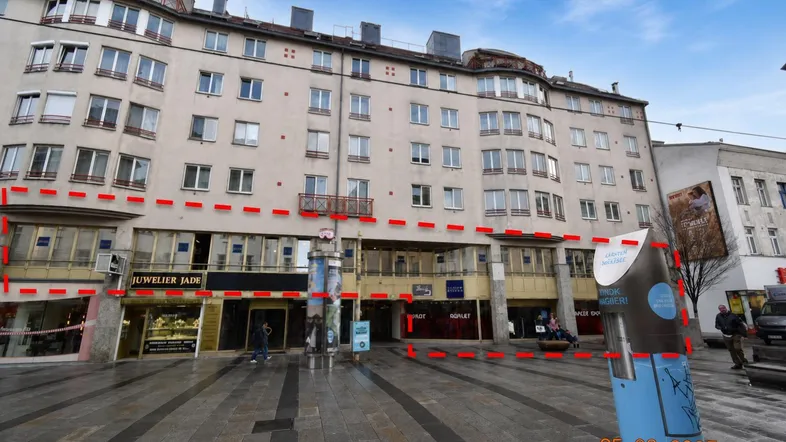 Gebäudeansicht mit Geschäften im Erdgeschoss und einer belebten Straße davor, markiert mit roten Linien.