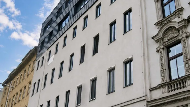 Moderne Gebäudefassade mit vielen Fenstern und angrenzendem Altbau unter blauem Himmel.