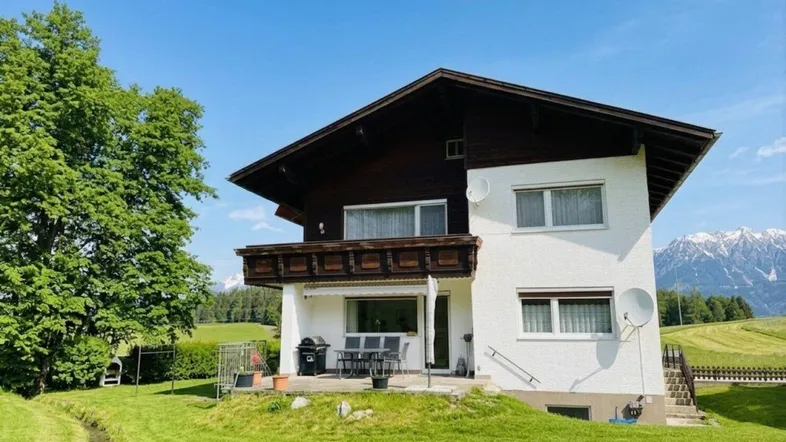 Haus im alpinen Stil mit Terrasse, Balkon und Garten, umgeben von grünen Wiesen und Bergblick.