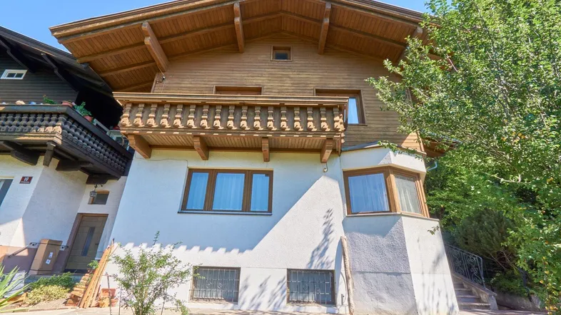 Traditionelles Holzhaus mit Balkon und weiß verputztem Untergeschoss, umgeben von viel Grün.