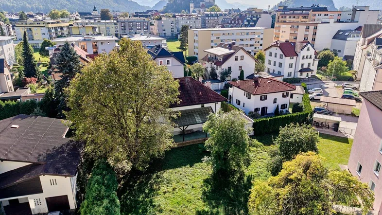 Luftaufnahme eines Grundstücks mit viel Grünfläche, umgeben von Wohngebäuden und Bergen im Hintergrund.