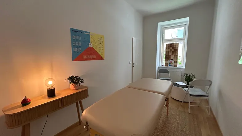 Heller Therapieraum mit Massageliege, Holztisch und Fenster, ideal für Behandlungen und Entspannung.