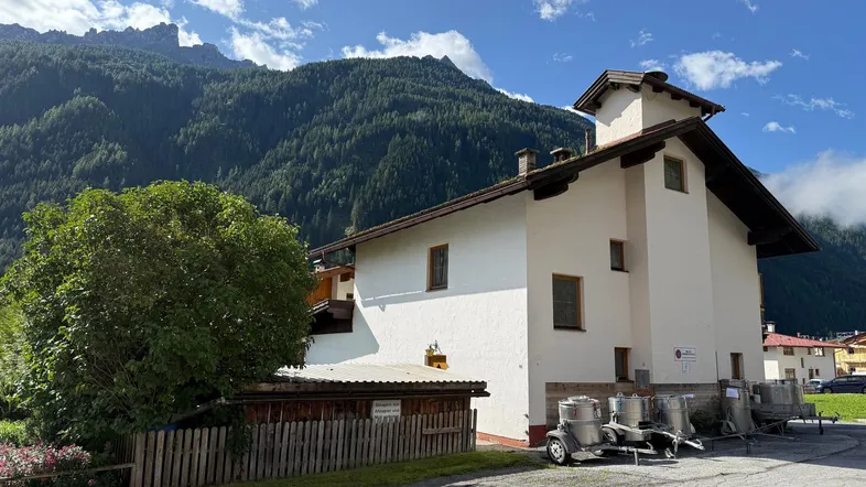 Weißes Gebäude mit braunem Dach und Bergblick, umgeben von grüner Natur.