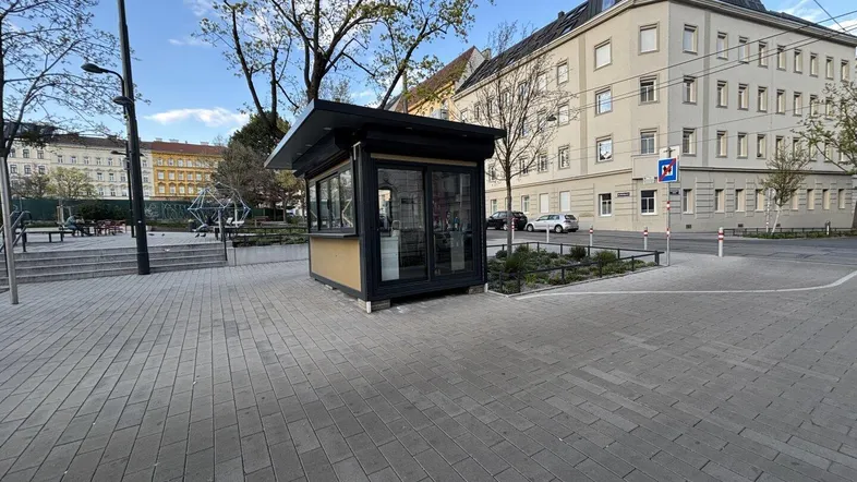 Kleiner, freistehender Kiosk auf einem gepflasterten Platz mit umliegenden Bäumen und Gebäuden.