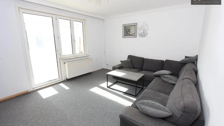 Helles Wohnzimmer mit grauem Teppichboden, Ecksofa und Zugang zu einem Balkon.