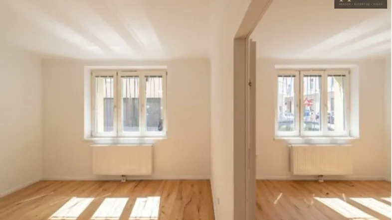 Helles, leeres Zimmer mit Holzboden und großen Fenstern, das viel natürliches Licht hereinlässt.