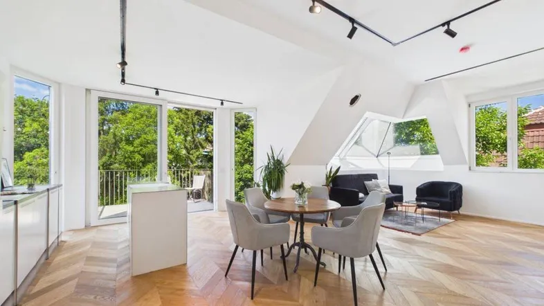 Heller, offener Wohn-Essbereich mit moderner Küche, Fischgrätparkett und Zugang zu einem Balkon mit Grünblick.