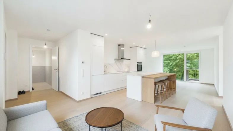 Heller, offener Wohn- und Essbereich mit moderner Einbauküche, Kücheninsel und Zugang zum Balkon mit Grünblick.
