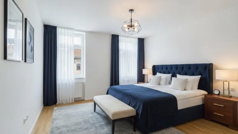 Helles Schlafzimmer mit großem blauen Bett, Holzboden und zwei Fenstern mit dunklen Vorhängen.