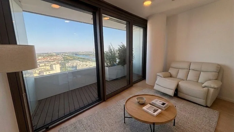Helles Wohnzimmer mit beigem Sofa, Holzboden und Zugang zu einem Balkon mit Stadtblick.