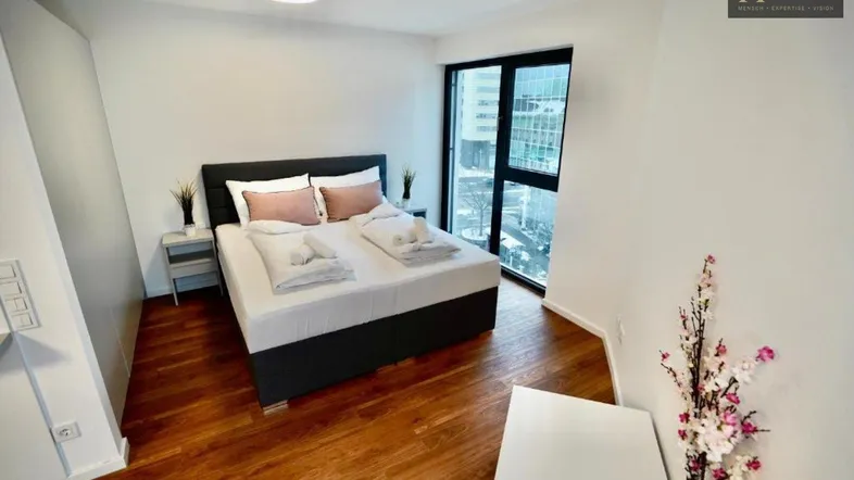 Helles Schlafzimmer mit Holzboden, Doppelbett und großem Fenster mit Stadtblick auf Hochhäuser.
