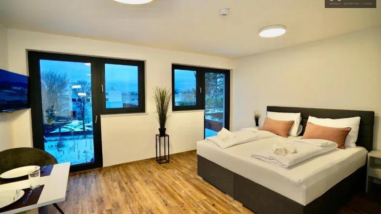 Helles Schlafzimmer mit großem Bett, Holzboden und Zugang zu einem Balkon oder einer Terrasse.