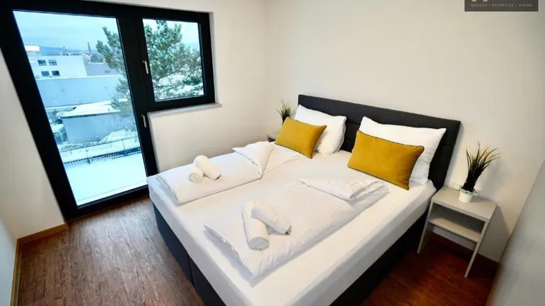 Helles Schlafzimmer mit Doppelbett, Holzboden und großem Fenster mit Blick ins Freie.