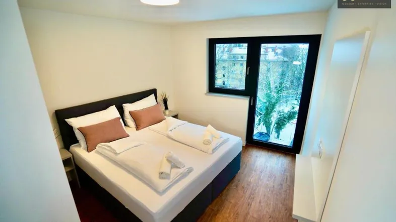 Helles Schlafzimmer mit Doppelbett, Holzboden und Zugang zu einem Balkon oder einer Terrasse.