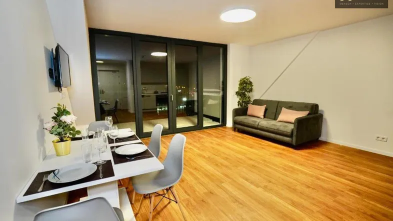 Helles Wohn- und Esszimmer mit Holzboden, Sofa und Zugang zu einem Balkon.