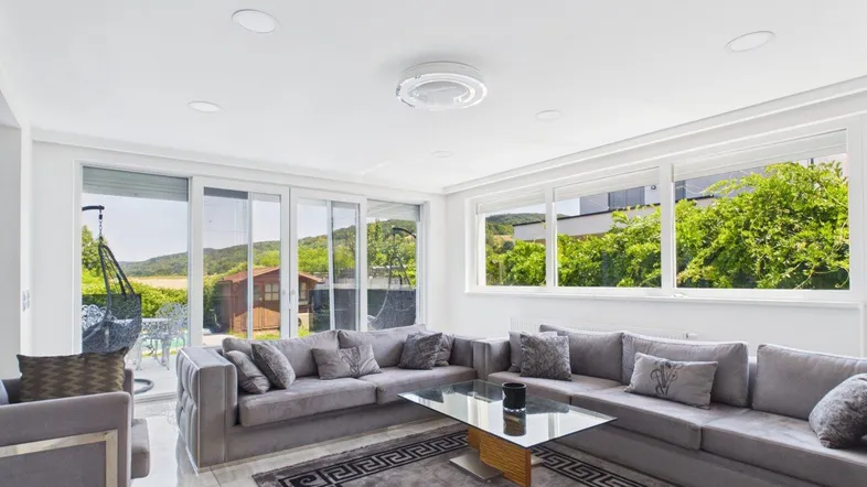 Helles Wohnzimmer mit großem Ecksofa, Fliesenboden und Zugang zur Terrasse mit Blick ins Grüne.