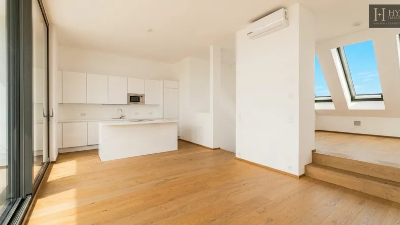 Heller offener Wohnbereich mit moderner Einbauküche, Holzboden und Zugang zu einem Balkon sowie Dachfenstern.