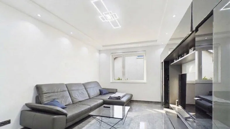 Modernes Wohnzimmer mit grauem Ecksofa, Fliesenboden und stilvoller Deckenbeleuchtung.