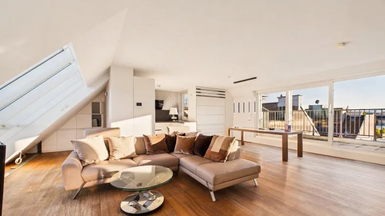 Helles Dachgeschoss-Wohnzimmer mit Holzboden, gemütlichem Sofa und Zugang zur Dachterrasse.