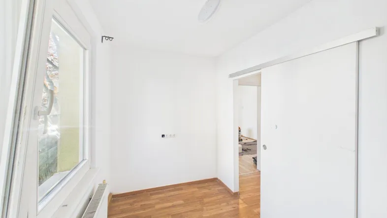 Heller Raum mit Holzfußboden und großem Fenster, der auf eine Renovierung wartet.