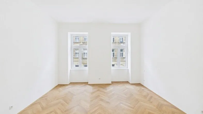Helles, leeres Zimmer mit Fischgrätparkett und zwei Fenstern, bereit zur individuellen Gestaltung.
