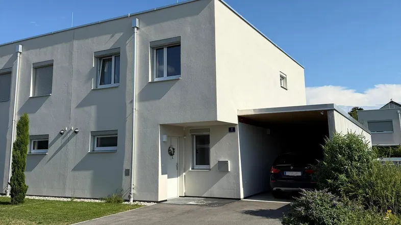 Modernes Reihenhaus mit heller Fassade, Vorgarten und Carport unter blauem Himmel.