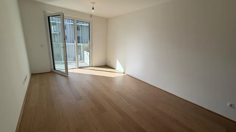 Helles, leeres Zimmer mit Holzboden und Zugang zu einem Balkon, durchflutet von natürlichem Sonnenlicht.