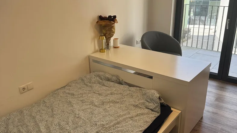 Helles Schlafzimmer mit Einzelbett, Schreibtisch und Zugang zu einem Balkon, ausgestattet mit Holzfußboden.